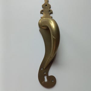 Puede incluir: Un pomo de puerta de latón con un agarre curvo y alargado y una placa decorativa con un remate ornamentado en la parte superior y una cerradura en la parte inferior. La superficie metálica muestra un acabado pulido con sutiles signos de desgaste.