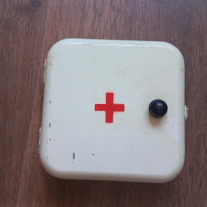 Op de afbeelding: Een vierkante, gebroken witte metalen verbanddoos met een opvallend rood kruis in het midden van de deur. De kast is voorzien van een kleine, ronde zwarte knop om de deur te openen en vertoont lichte gebruikssporen langs de randen.
