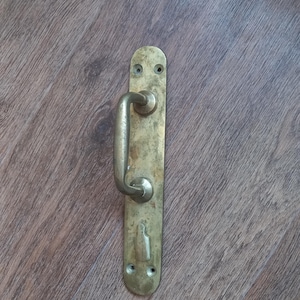 Peut inclure: Une poignée de porte en laiton patiné dotée d'une longue plaque rectangulaire aux extrémités arrondies. L'ensemble comprend une poignée incurvée et une ouverture pour serrure, avec deux trous de vis visibles en haut et deux en bas.