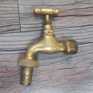 Puede incluir: Un grifo de latón con un mango en forma de T y un caño curvo. El grifo tiene un extremo roscado para la conexión y una sección acanalada para una manguera. El accesorio es de metal brillante de color dorado.