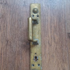 Puede incluir: Un tirador de puerta y placa de latón antiguos, con una placa trasera rectangular y un tirador curvo. El tirador está montado sobre una superficie de madera.