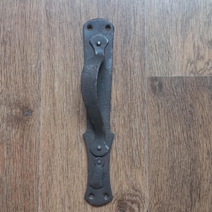 May include: A dark grey, wrought iron door handle with a curved grip. The handle is mounted on a rectangular plate with decorative detailing. The hardware is likely for a rustic or vintage-style door. The handle is designed for easy gripping and pulling.