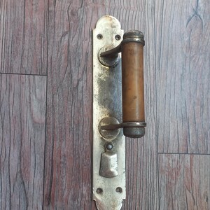 May include: A vertical metal door handle assembly featuring a weathered silver-toned rectangular backplate with rounded ends and four screw holes. A cylindrical brown handle is mounted to the plate, accompanied by a small, hinged metal latch below.