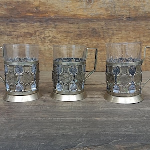 May include: Three vintage glass tea cup holders with ornate metal filigree designs. Each cup holder has a handle and sits on a round base. The cups are clear, and the metal has a gold tone. The background is a wooden surface.