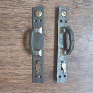 Puede incluir: Dos manijas de puerta de metal antiguas con un acabado envejecido. Cada manija tiene una placa trasera rectangular gris oscuro con una manija curva, detalles en latón y un ojo de cerradura. Las manijas están montadas sobre una superficie de madera.