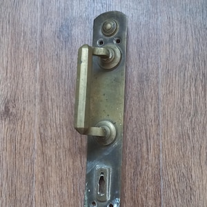 Op de afbeelding: Een messing deurkrukset met een rechthoekige achterplaat met afgeronde uiteinden, een zeshoekige handgreep en een sleutelgat. Het metalen oppervlak vertoont tekenen van ouderdom en oxidatie, met zichtbare schroefgaten aan de boven- en onderkant.