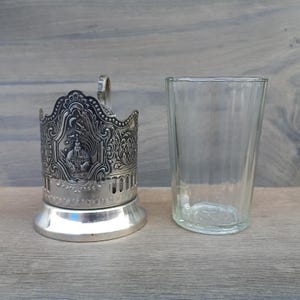 Puede incluir: Un portavasos de metal plateado ornamentado con un diseño en relieve detallado, junto a un vaso transparente y acanalado. El soporte presenta un asa y una base decorativa. El vaso está vacío.
