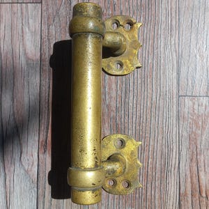 Op de afbeelding: Een cilindrische deurklink van messing met een verweerde, goudkleurige afwerking. De klink heeft twee decoratieve montageplaten aan de boven- en onderkant, elk met twee ronde schroefgaten en een gekartelde, geschulpte rand aan de buitenkant.