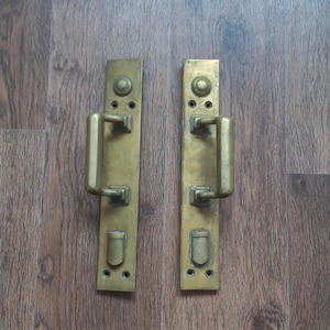 Könnte beinhalten: Zwei antike Türgriffe aus Messing. Jeder Griff hat eine rechteckige Rückplatte mit einem gebogenen Griff und einem kleinen, abgerundeten Detail oben und unten. Das Messing hat eine warme, gealterte Patina, die auf Vintage-Handwerkskunst hindeutet.