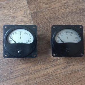 May include: Two vintage black analog meters with white faces and black needles. One meter displays "A" for amps, the other displays "V" for volts. Both are square with rounded dials and screw holes.