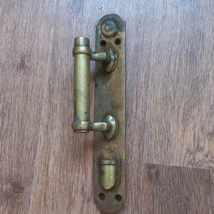 May include: A vintage brass door handle assembly featuring a long rectangular backplate with rounded ends, a vertical cylindrical grip, and a lower latch mechanism. The metal surface shows signs of age, oxidation, and a weathered patina throughout.