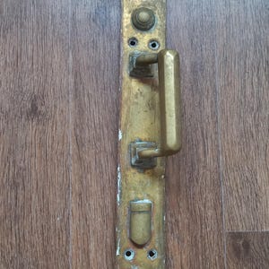 Könnte beinhalten: Ein antiker Türgriff und eine Platte aus Messing, die Gebrauchsspuren aufweisen. Der Griff hat ein einfaches, rechteckiges Design, und die Platte ist vertikal ausgerichtet mit Befestigungslöchern und dekorativen Elementen. Die Hardware ist an einer Holztür montiert.