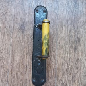 May include: A vertical door handle featuring a long, black metal backplate with rounded ends and a diamond-shaped embossed detail. Attached to the plate is a cylindrical handle grip with a yellow and black marbled finish and metal end caps.