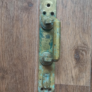 Puede incluir: Un tirador de puerta antiguo de latón con placa trasera rectangular. El tirador tiene un agarre vertical y rectangular y pomos circulares. El latón tiene un aspecto envejecido con toques de pátina azul-verdosa, montado sobre una superficie de madera.