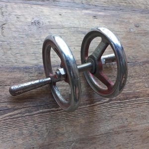 Pode incluir: Um volante industrial vintage com acabamento prateado e detalhes em vermelho. O volante tem uma haste roscada central e é montado em uma superfície de madeira. A alça é cilíndrica e o volante tem um design clássico e funcional.