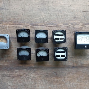 Puede incluir: Ocho medidores analógicos vintage con biseles negros y caras blancas. Los medidores están dispuestos en dos filas de cuatro. Los medidores están sobre una superficie de madera.
