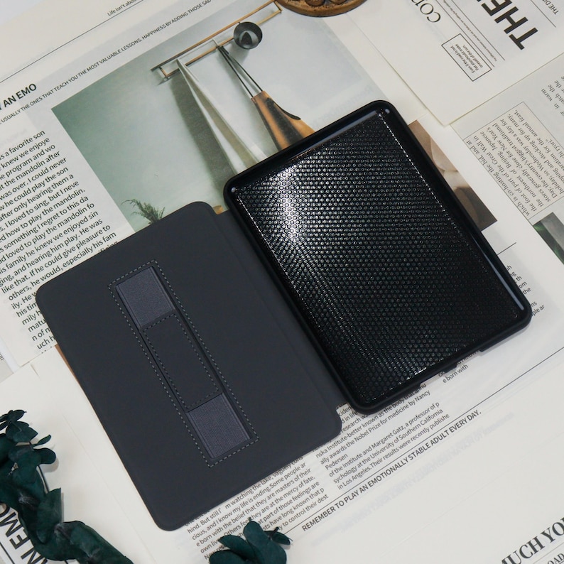 K&ouml;nnte beinhalten: Eine schwarze Tableth&uuml;lle mit strukturierter Oberfl&auml;che und dunkelgrauem Innenraum. Die H&uuml;lle ist ge&ouml;ffnet und zeigt den Tablet-Bildschirm. Die H&uuml;lle hat einen eingebauten St&auml;nder f&uuml;r freih&auml;ndiges Betrachten. Der Hintergrund enth&auml;lt Zeitungsausschnitte.