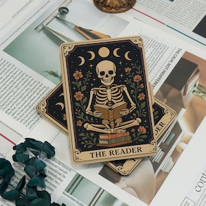 May include: Tarot card featuring a skeleton reading a book, surrounded by flowers and crescent moons. The card has a black background with the words "THE READER" at the bottom. The card is beige with a decorative border.