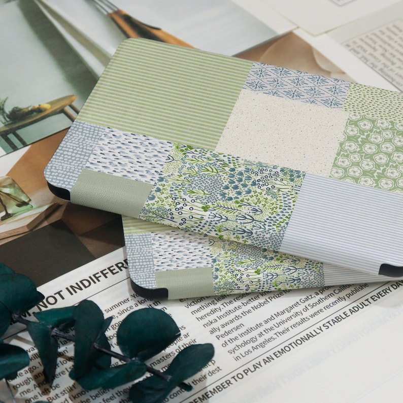 May include: Two notebooks with a patchwork design featuring various green and blue patterns. The notebooks have a soft, muted color palette and are resting on a background of printed papers and a sprig of green leaves.