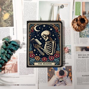 May include: A rectangular tarot card depicting a skeleton reading a book beneath a crescent moon and stars. The card has a dark blue background with floral and star accents. The text "THE READER" is at the bottom.