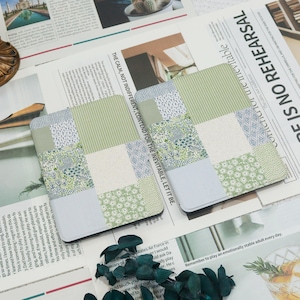 May include: Two square coasters with a patchwork design. Each coaster features various patterns, including stripes, floral prints, and geometric shapes, in shades of green, blue, and white. The coasters are placed on a newspaper background.