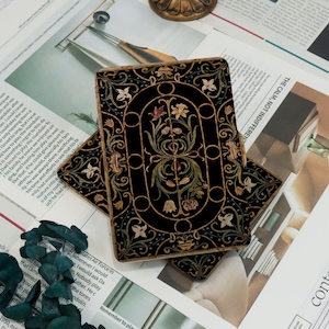 May include: Two decorative black coasters with intricate floral designs in gold, beige, and green. The coasters have a gold border and are placed on a newspaper background. The coasters are likely for home decor.