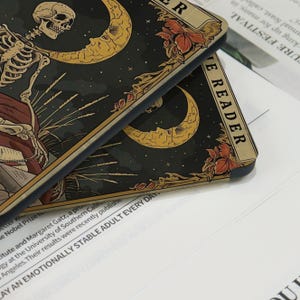 May include: Two phone cases with a tarot card design. The cases have a dark background with a skeleton, crescent moons, and floral accents. The text "THE READER" is visible on one case.
