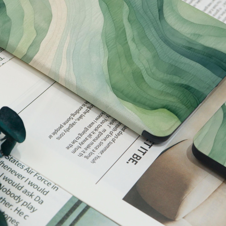 K&ouml;nnte beinhalten: Nahaufnahme eines dekorativen Gegenstands mit Aquarelldesign. Das Objekt zeigt ein geschichtetes Muster aus gr&uuml;nen und wei&szlig;en Farbt&ouml;nen, das an H&uuml;gel oder Wellen erinnert. Die R&auml;nder sind dunkel, und es liegt auf einer Zeitung.