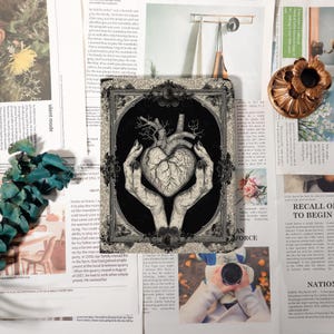 May include: Black and white illustration of a heart with tree-like veins held by two hands, within a decorative frame. The image is on a tablet or similar device, surrounded by newspaper clippings and other decorative items.