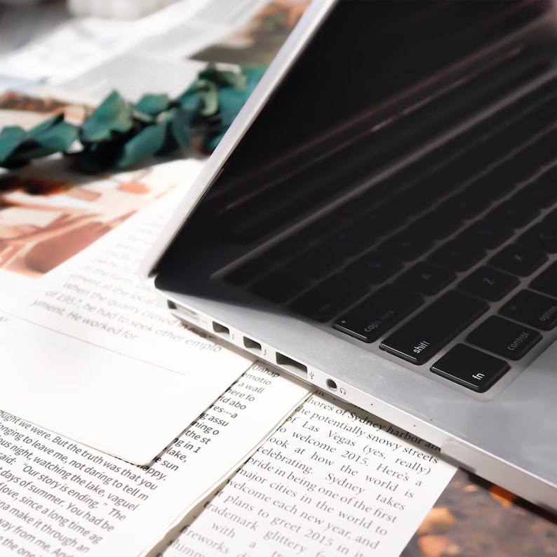 May include: A silver laptop computer with a black keyboard rests on top of printed pages. The screen is dark, and the keyboard is in focus. The pages have text and are partially visible. Green leaves are in the background.