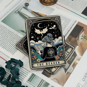 May include: Two tarot cards featuring a woman reading under a night sky with dragons and a crescent moon. The cards have a black border with floral accents and the text "THE READER".