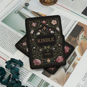 May include: Black Kindle cover with floral design, the word "KINDLE" in a circular frame, and the name "SOPHIA" below. The cover features pink and white flowers, green leaves, and a cross. The background includes newspaper clippings.