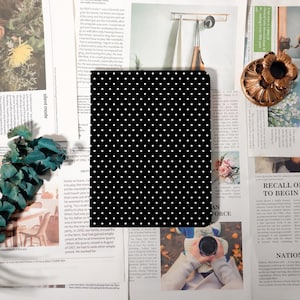 May include: A black notebook with a white polka dot pattern. The notebook is rectangular and appears to be closed. The background includes newspaper clippings and decorative items.
