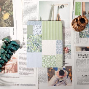 May include: A square, patchwork journal with various patterns in green, blue, and white. The patterns include stripes, floral designs, and geometric shapes. The journal is surrounded by newspaper clippings and decorative elements.