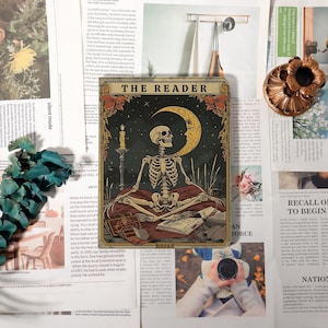 May include: A decorative square metal sign featuring a skeleton meditating in front of a crescent moon and a lit candle. The sign has the words "THE READER" at the top and "BOOKS" at the bottom. The color palette includes gold, brown, and black.