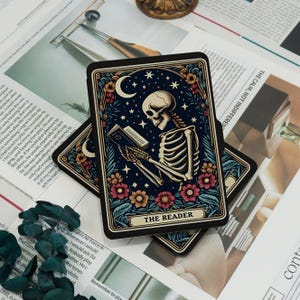 May include: A tarot card featuring a skeleton reading a book under a crescent moon and stars. The card is framed by flowers and foliage, with the text "THE READER" at the bottom. The card has a dark blue background.