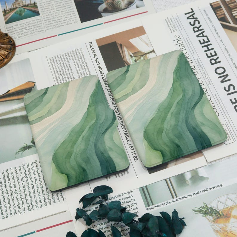 K&ouml;nnte beinhalten: Zwei quadratische Untersetzer mit einem Aquarelldesign aus geschichteten gr&uuml;nen und wei&szlig;en Wellenlinien. Die Untersetzer befinden sich auf einem Zeitungs-Hintergrund mit Text. Ein Einrichtungsgegenstand.