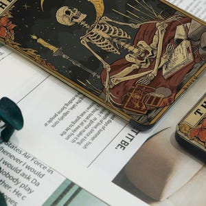 May include: A tarot card depicting a skeleton resting on a burgundy cushion, surrounded by books and a candle. The card has a dark background with a crescent moon and stars. Part of a tarot deck.