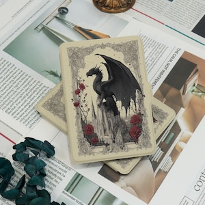May include: A rectangular beige case with a black dragon design, set against a gothic castle and red roses. The dragon has large wings and is set against a detailed, vintage-style background. The case is likely for a phone or tablet.