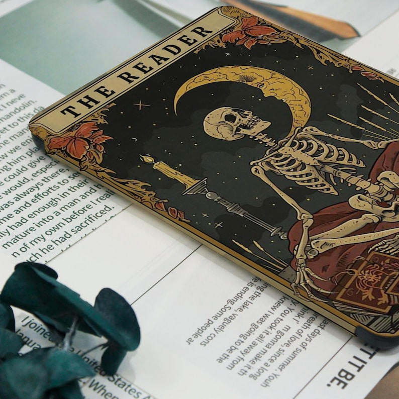 May include: A rectangular tarot card with a skeleton, crescent moon, stars, and a candle. The card is labelled "THE READER". The colour scheme includes black, gold, red, and beige.