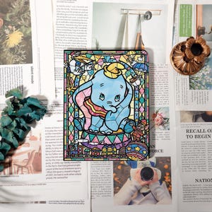Peut inclure: Une œuvre d'art colorée de style vitrail représentant Dumbo l'éléphant. L'œuvre présente un éléphant bleu avec un chapeau jaune, entouré d'éléments sur le thème du cirque et d'une bordure décorative. L'image est posée sur un fond de coupures de journaux.