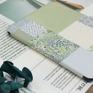 May include: A patchwork-style phone case featuring various patterns in shades of green, blue, and white. The case is made of fabric and has a rectangular shape. The design includes floral, striped, and dotted patterns. The case is lying on a printed paper.