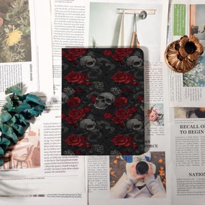 Könnte beinhalten: Ein schwarzes Notizbuch mit einem sich wiederholenden Muster aus grauen Totenköpfen, roten Rosen und Würfeln. Das Notizbuch ist von Zeitungsausschnitten und anderen dekorativen Elementen umgeben, was eine gotische Ästhetik erzeugt.