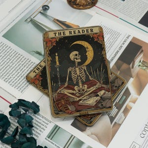 May include: Tarot card featuring a skeleton meditating with a crescent moon and candle. The card is titled "THE READER" and has a dark, celestial background with floral accents. Books are visible at the bottom of the card.