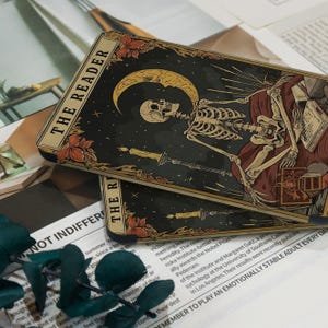 May include: Two tarot cards titled "THE READER" are shown. The cards depict a skeleton figure with a crescent moon, candles, and floral details. The background is dark with stars.