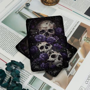 May include: Two black coasters with a skull and purple rose design. The skulls are white with dark shadows, and the roses are a deep purple. The coasters are stacked on a newspaper background.