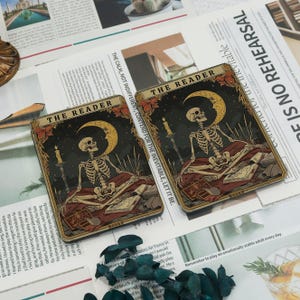 May include: Two tarot cards depicting a skeleton figure seated with a book, a crescent moon, and candles against a starry night sky. The cards have the text "THE READER" and "BOOKS". The cards are on a background of printed paper.