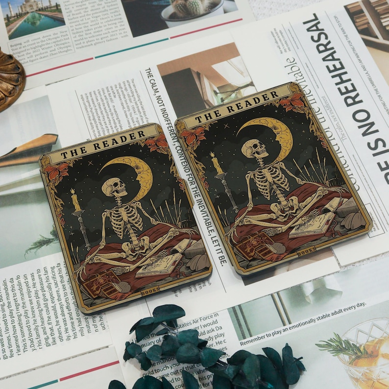 May include: Two tarot cards depicting a skeleton figure seated with a book, a crescent moon, and candles against a starry night sky. The cards have the text "THE READER" and "BOOKS". The cards are on a background of printed paper.