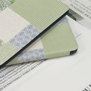 May include: Close-up of two notebooks with a patchwork design. The covers feature various patterns, including floral, striped, and dotted designs in shades of green, blue, and cream. The notebooks have black edges and are resting on a white surface with text.