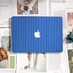 May include: Blue laptop with a striped pattern and a white Apple logo. The design features alternating light and dark blue vertical stripes. The laptop is on a newspaper background.